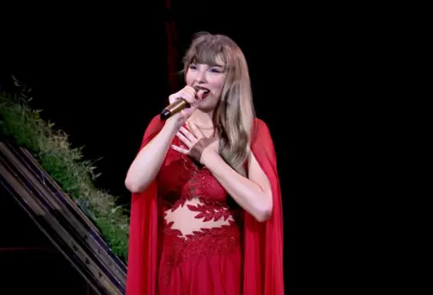 Getty Images Taylor Swift onstage in a red dress and cape, during the Folklore/Evermore set of her Eras Tour