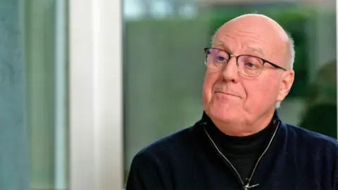 Stanley wears glasses and a navy top and looks serious