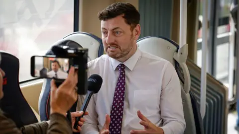 Getty Images Cammy day in a shirt and tie sitting on a tram seat being interviewed by a woman holding up an iphone and a microphone