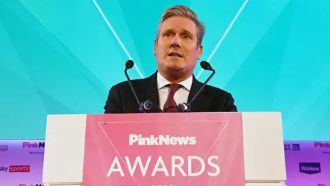 Getty Images Sir Keir Starmer standing at a lectern with a sign reading "PinkNews Awards", wearing a  dark suit and tie, with a turquoise background behind him. 