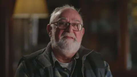 Journalist Andy Spearman looks into the camera through his glasses in a dark lit room, with the background out of focus. He has grey thinning hair and a short grey beard. Andy is wearing a white t-shirt, green and navy checked shirt and a navy body warmer. 