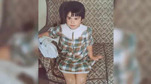 Rebecca Rebecca aged about four years old sits on an old fashioned looking sofa, her arm over the arm rest. She is wearing a green and cream tartan dress which has a big white collar. The photo is from the 1980s. Rebecca has short dark hair, freckles on her nose and is looking past the camera at someone else in the room. 