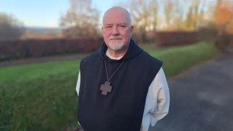 Father Jan Rossey stands in a traditional monk's habit - white robe covered in a black hooded robe with a cross on a long chain around his neck. His hands are behind his back and he's looking into camera, smiling slightly. He is bald with a white goatie beard.