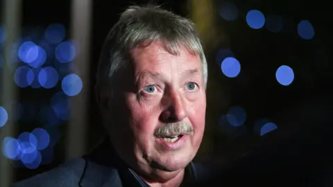 Reuters Sammy Wilson with spotlights in the background. He has short grey-ish hair, blue eyes and a moustache and wearing a dark grey blazer. 