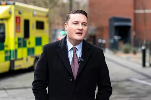 PA Media Health Secretary Wes Streeting is seen speaking to reporters outside a hospital. He is wearing a black coat and purple tie. Over his right shoulder an ambulance is seen parked.