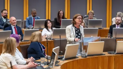 Welsh Government/PA Eluned Morgan wearing a white jacket and standing, addresses the Senedd with other Labour politicians listening 