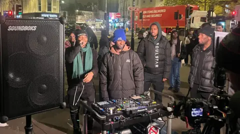 DJ AG wearing a black jacket and a blue bobble hat behind decks and a big speaker looking into a camera as he streams to thousands of people. Someone's rapping next to him wearing all black with a green scarf. 