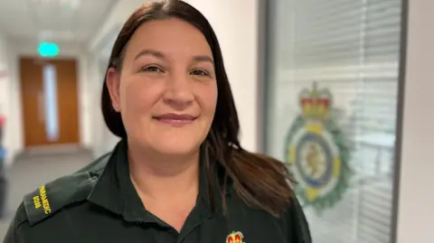 Amy is wearing a green paramedic uniform and has long, dark hair. She is stood in an office environment with the ambulance service logo on an office window in the background.