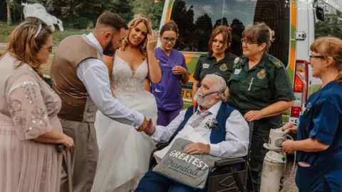 CJ Photography Wales Wayne is in a wheelchair, with an oxygen mask, wearing a suit at his daughter's wedding. He is surrounded by two paramedics and two nurses, next to an ambulance. He is shaking the hand of his son-in-law, next to his daughter, who is wearing a wedding dress. His wife is also in the picture.