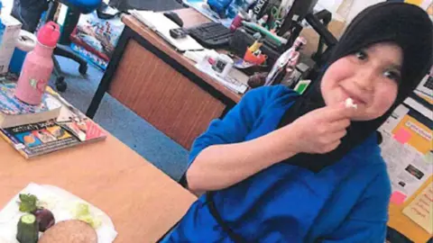 Sara Sharif wearing a hijab and eating a school dinner in a classroom. She is smiling and holding up a piece of food to her mouth.