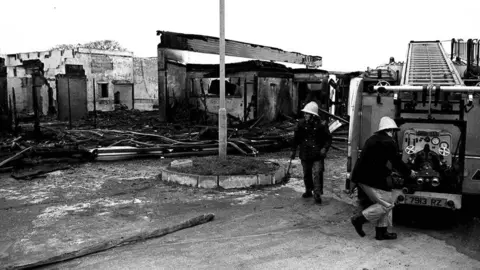 Pacemaker Black and white image of the bombed La Mon hotel, the brick building is covered in black with parts with most of the building frame collapsed, debris lies on the floor and two men stand by an old fire engine outside the building they are wearing dark jackets and trousers with black wellington boots and  hard hats. One man is getting the hose from the back of the fire engine.