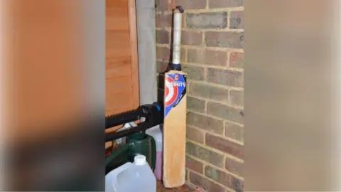 Surrey Police A cricket bat found near an outhouse at the family's home in Woking, It is wooden and has a white handle, with bricks in the background.