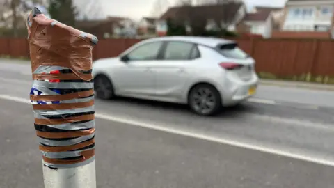 A stretch of road, in the forefront of the picture and to the left is a taped up broken camera, there is a silver car driving on the road.