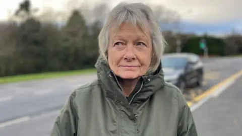 A woman on the side of the road, behind her is a black car driving, she is the focus of the image. She is looking at the camera, she has grey hair and is wearing a khaki cagoule. 