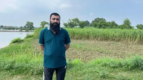 Mr Munir dressed in a blue T-shirt and black tracksuit pants stands next to a field of corn which is next to a body of water.