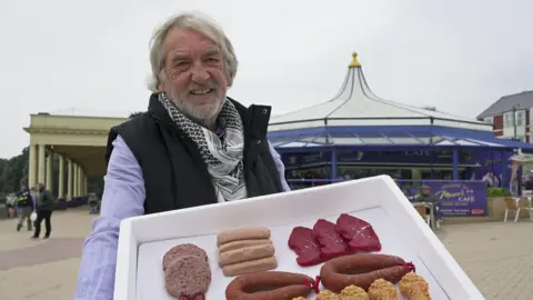 Gwynfor Roberts poses for a photo on Barry Island seafront holding barbecue meats, recreating his scene from series three