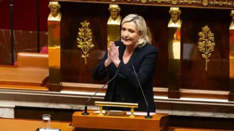 Getty Images Marine Le Pen, France's far-right National Rally leader, addresses the National Assembly wearing a dark-coloured jacket and holding her hands together