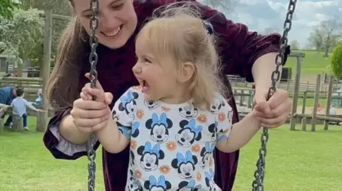 Facebook Isabella smiling on a swing with her mother smiling and standing behind her. Isabella has a T-shirt on with Minnie Mouse's face on it.
