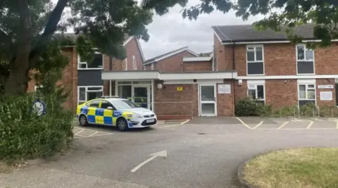 Ben Parker/BBC A police car parked outside a red-brick, double-storey housing complex. There is a car park to the front of the building and a green patch of grass.
