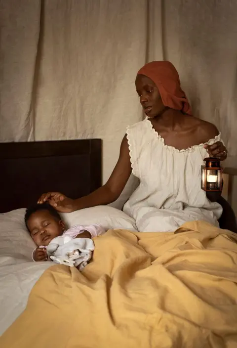 Dola Posh Dola Posh wears an orange headscarf and a white nightie. She is holding a lamp and standing by a bed where her daughter is sleeping.