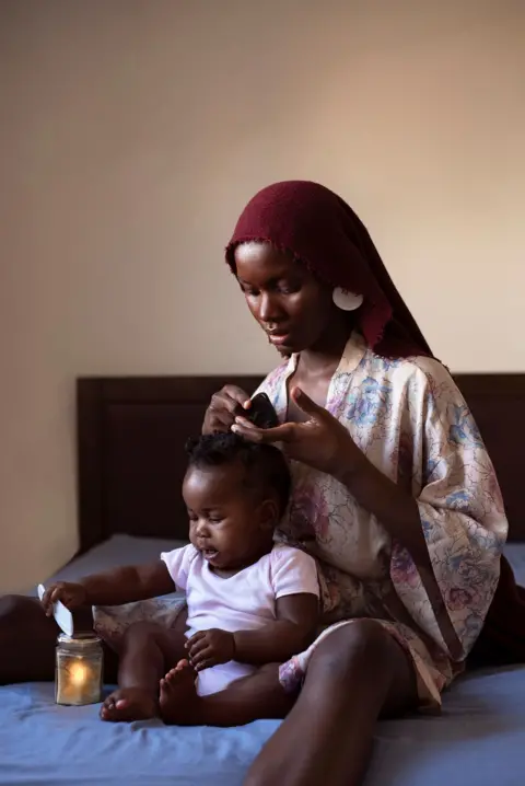 Dola Posh Dola Posh, with a red head covering, combs her daughter's hair as they sit on a bed with a blue covering. There is a candle in a jar next to them.