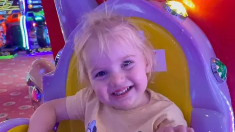 PA Isabella Jonas-Wheildon, a young blonde toddler wearing a yellow t-shirt smiling at the camera. She is sitting on a yellow and purple chair at an arcade.
