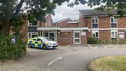 Ben Parker/BBC A police car parked outside a red-bricked building. The building has a car park to its front.
