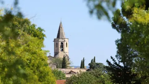 Reuters A church spire and houses seen from between green trees and undergrowth, set against a bright blue sky