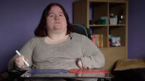 John Boon/BBC Emma in her living room which is decorated purple. Emma is sitting on her electric wheelchair looking into the camera