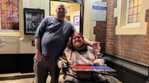 Handout Emma and Wes are outside their local pub. Emma is in her electric wheelchair while Wes is standing next to her on the left of the picture.