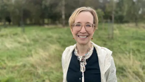 Louise McManus has short blonde hair. She wears round glasses, a navy cardigan with a blouse tied in a bow at the neck, and a cream coloured waterproof jacket. She is smiling broadly at the camera and standing in a field. There are a few young trees behind her.