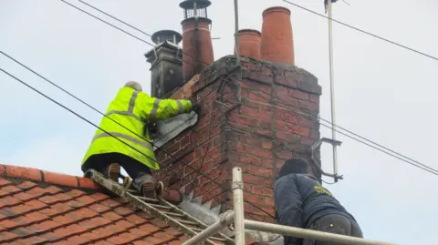 Angela Clarke Two men, one wearing a hi-vis jacket, are on a roof. One is fixing a panel to the chimney. Scaffolding has been put up around the house and there is a ladder laid over the roof.