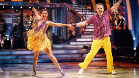 BBC/PA Diane Buswell wears a yellow and purple tasselled dress and Chris McCausland is pictured in a pink shirt and yellow trousers on the Strictly Come Dancing dancefloor.