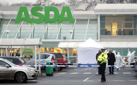 PA Media A lone police officer stands guard in front of blue and white police tape in the car park of Asda, Robroyston. Behind the tape a white forensic tent has been erected and specialists in white hazmat suits can be seen carrying out inquiries. Plain clothes detectives, including one wearing black trousers and a long black coat, are also visible in the background.