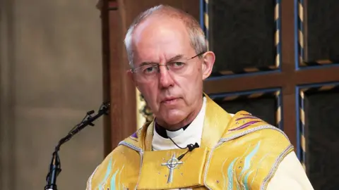 Getty Images Archbishop of Canterbury Justin Welby pictured in front of a small microphone in a church pulpit. He has receding short grey hair, rimless glasses and is clean shaven. He is wearing gold coloured robes embellished with purple and turquoise waves and jewels, and a silver embroidered crucifix. 