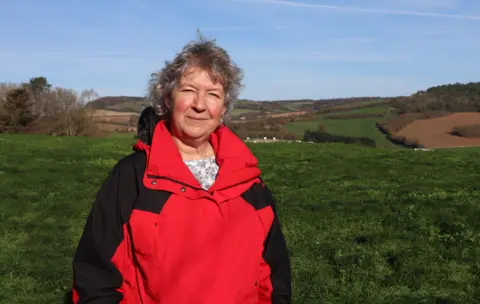 Steve Swann / BBC Portrait picture of former vicar Jenny Penn. She has short, curly, grey hair and is wearing a patterned t-shirt under a red and black hooded waterproof jacket. She is pictured outdoors with blue skies and green fields behind her. 