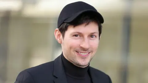 Getty Images Pavel Durov, who has brown hair, brown eyes and light stubble and is wearing a black suit, a black T-shirt and a black baseball cap, smiling at the camera outside a Paris court for a hearing