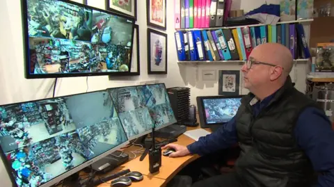 Martin Gaunt wearing his blue shirt under black gillet looking at three large CCTV camera screens which are spilt into four showing differing views. He is wearing glasses looking at the screens and sitting at a desk holding a mouse. Shelves containing colourful ring binders and another screen are in the background.