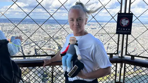 Family photo Alex standing on the Eiffel Tower viewing platform, holding a knitted version of Declan