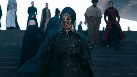 Attila Szvacsek/HBO Tabu, playing the character of Francesca, dressed in all black, with a netted face veil, staring into the distance, while standing on dark coloured steps with people walking in the background.