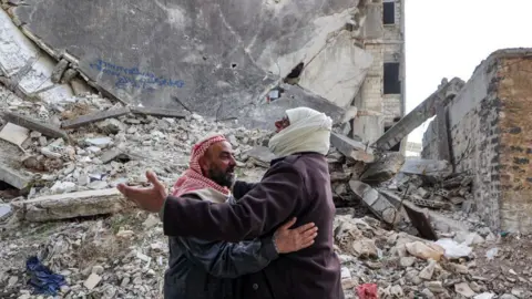 Getty Images Two men in scarves embrace in front of a building turned into rubble in Baba Amr district of Homs on 6 December