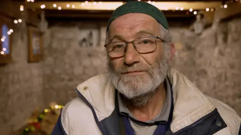 Francesco Tosto/BBC Farhan Abdul Ghani stands in front of camera wearing a hat and glasses, and a white beard, inside a stone-walled room