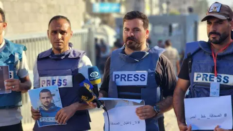 Getty Images Four Palestinian journalists are pictured, all of them wearing blue flak jackets with the word "press" on them. They are holding pictures of Hamza Murteca in his memory. It appears to be a sunny day and they are all standing on a street in Gaza.