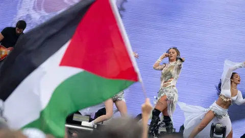 Getty Images Elyanna performing on stage in San Francisco. She is wearing a white and silver glittery outfit, with her right hand going towards her right ear and her left hand is holding a black microphone outstretched towards the crowd. In the crowd, a fan is holding the flag of Palestine, which is coloured red, black, white and green.