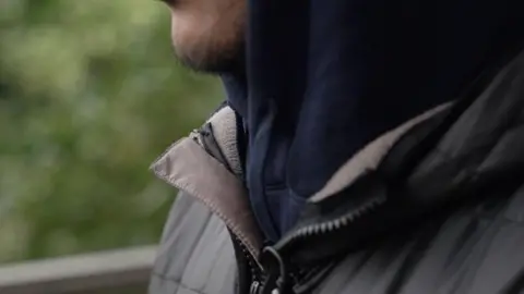 A person wearing a dark jacket shown against a soft-focus outdoor background. He appears to be photographed from chest up, shown in natural lighting with an urban setting behind him.