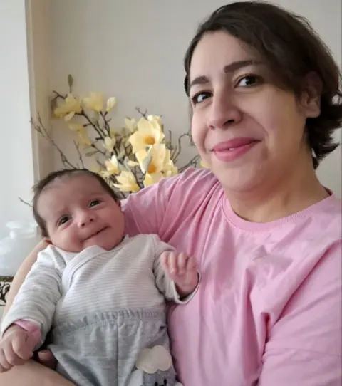 UGC Photograph of Rezvaneh holding her baby daughter. Rezvaneh has short to medium-length dark hair, is smiling and is wearing a light pink top. Her daughter has dark hair and is wearing white and grey striped top and grey trousers. There are yellow flowers in the background.