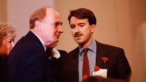 Getty Images Then Labour leader Neil Kinnock confers with a moustachioed Peter Mandelson in 1989. Both men are wearing dark suits