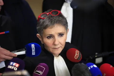 EPA Béatrice Zavarro with grey hair and red-rimmed glasses on her head stands in front of several microphones as she speaks to the press.