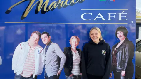 Ellie, who has blonde hair and is wearing a black hoodie, standing in front of murals of the cast outside Marco's Cafe.