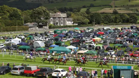 Getty Images Cartmel Racecourse pictured during the Hadwins Motor Group Handicap Hurdle race in 2017. Horses are racing and the course is surrounded by cars and gazebos.